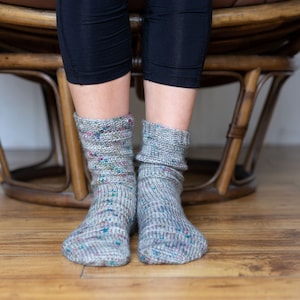 May include: A pair of grey socks with a speckled pattern. The socks are worn by a person sitting in a wicker chair.