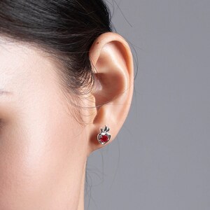 Puede incluir: Primer plano de la oreja de una mujer con un pequeño pendiente en forma de corazón rojo con un diseño de llama plateado.