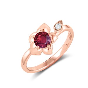 Op de afbeelding: Een ring van roségoud met een rode edelsteen die in een bloemvorm is gezet. Een kleinere diamant is aan de zijkant van de ring gezet.