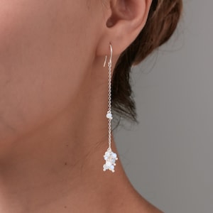 May include: A delicate silver earring with a long chain and a cluster of small, white, iridescent beads at the bottom. The earring is shown worn on an ear, with a simple hook design.
