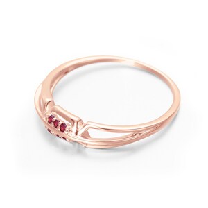 May include: A delicate rose gold ring with a criss-cross design and a row of small red gemstones.