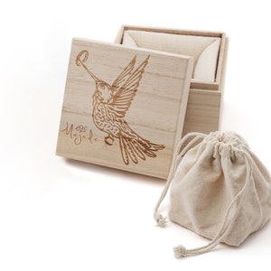 May include: A wooden box with a hinged lid, featuring a laser-engraved hummingbird design and the word "Magdal" on the lid. The box is open, revealing a beige fabric lining. A small, beige drawstring bag is also included.