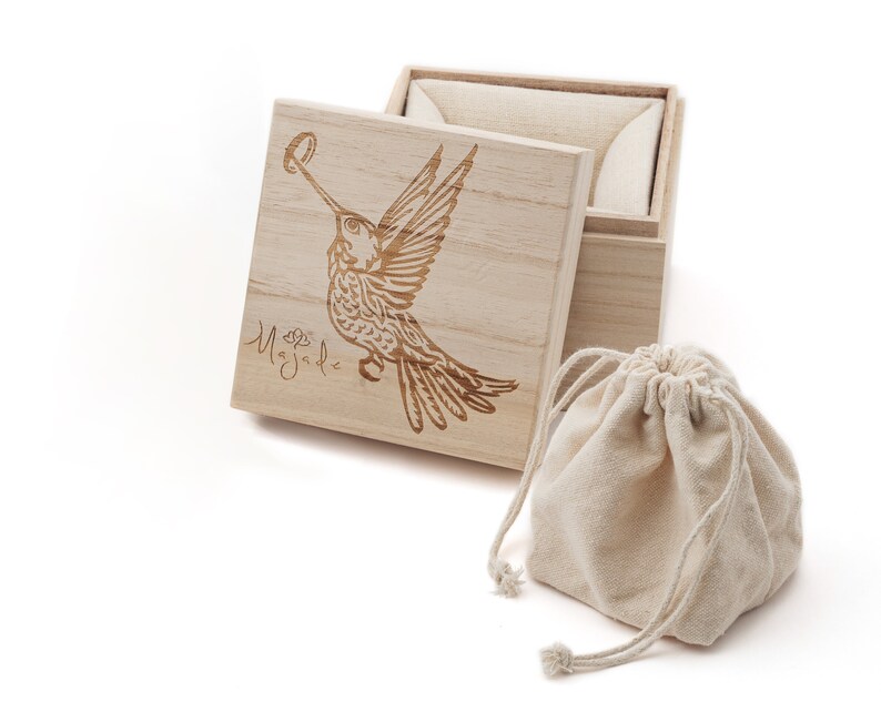 May include: A wooden box with a hinged lid, featuring a laser-engraved hummingbird design and the word "Magdal" on the lid. The box is open, revealing a beige fabric lining. A small, beige drawstring bag is also included.