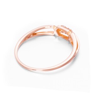 May include: A rose gold ring with a delicate, intertwined band design. The ring features a small, clear gemstone set in a square-shaped setting.