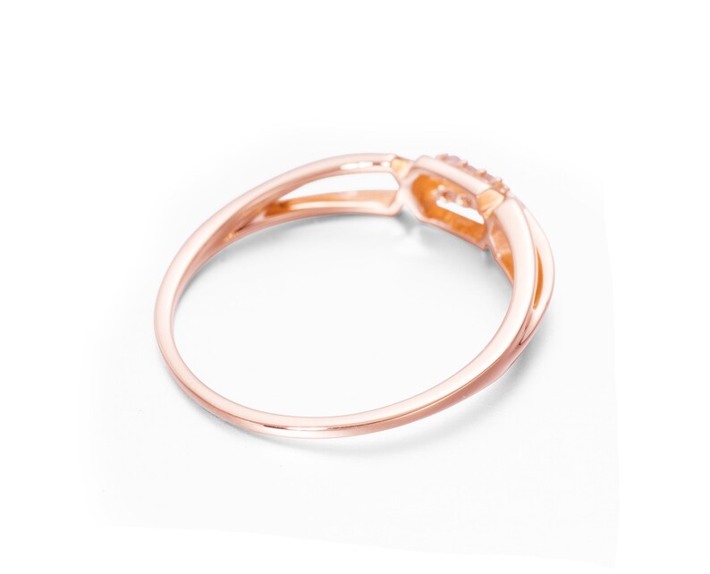 May include: A rose gold ring with a delicate, intertwined band design. The ring features a small, clear gemstone set in a square-shaped setting.