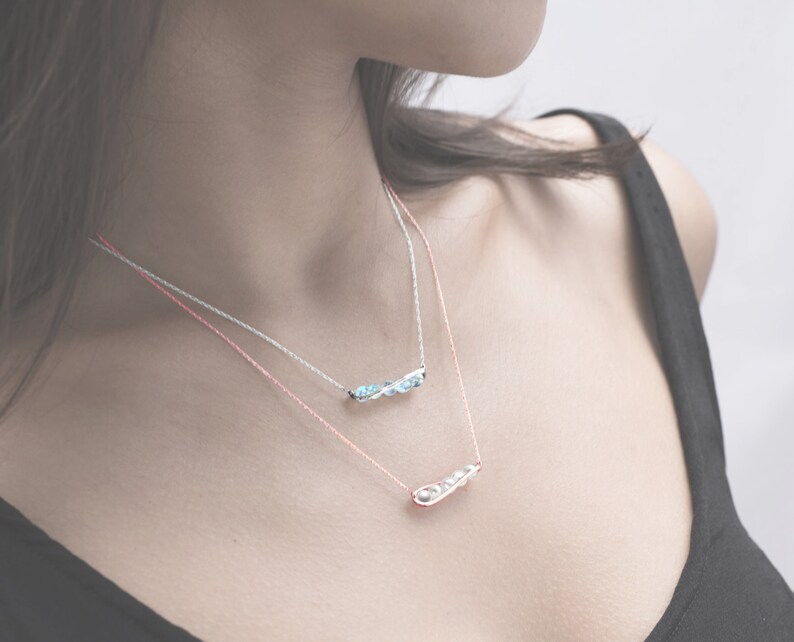 May include: Two delicate necklaces with silver chains and small charms. The top necklace features a row of light blue stones, while the bottom necklace has a single charm shaped like a pea pod.
