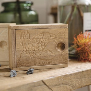 May include: A wooden box with a carved fish design and the word "MAJADE" on the front. The box is open and has a small compartment inside.