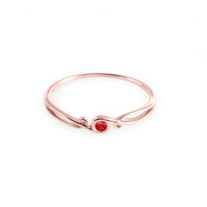 May include: A delicate rose gold ring with a single red gemstone set in the centre. The ring features a unique, flowing design.