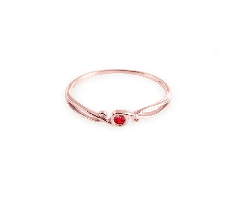 May include: A delicate rose gold ring with a single red gemstone set in the centre. The ring features a unique, flowing design.