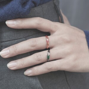 May include: Two rings on a finger. The top ring is gold with red stones. The bottom ring is black with green stones.