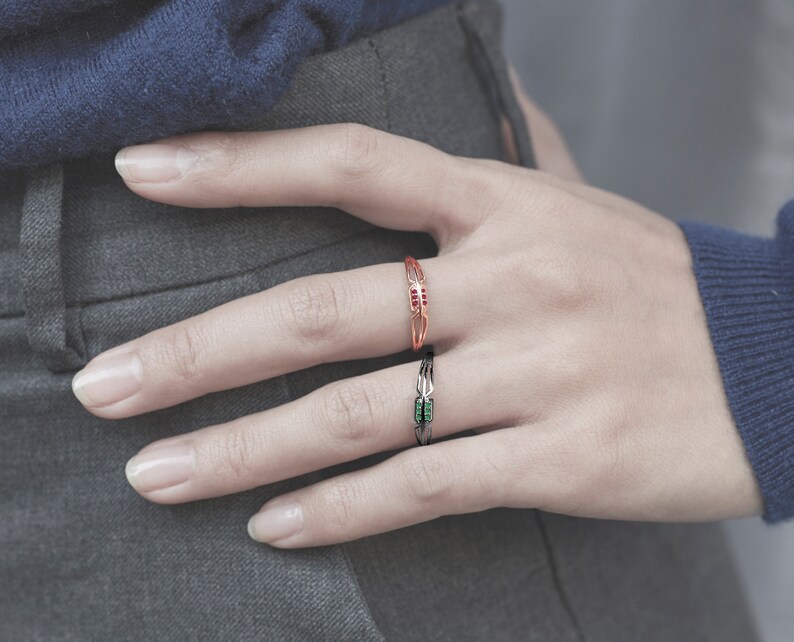 May include: Two rings on a finger. The top ring is gold with red stones. The bottom ring is black with green stones.