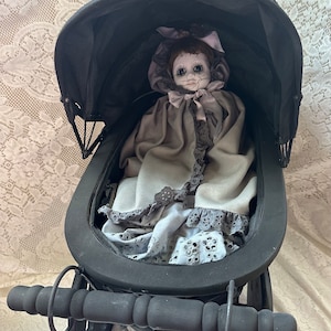 May include: A black doll stroller with a doll wearing a grey dress and a pink bonnet. The doll has a painted face with large eyes and a cracked appearance.