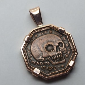 May include: A gold-toned pendant featuring a detailed skull with a floral design and the inscription "Memento Vivere".