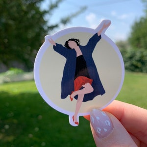 May include: A round sticker featuring a stylized illustration of a woman dancing. She wears a navy blue coat, a black dress, and a red skirt and shoes. The sticker has a cream-colored background and is held by a hand with a light pink nail polish.