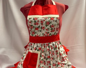 May include: A red and white floral apron with a ruffled hem and a large pocket. The apron has a white trim and a red bow at the neck.