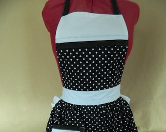 May include: A black and white polka dot apron with a white ruffle trim and a white pocket. The apron has a white top section and a black band.