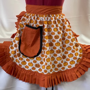 Puede incluir: Un delantal naranja y blanco con estampado de calabaza y un gran bolsillo naranja. El delantal tiene un borde inferior con volantes.