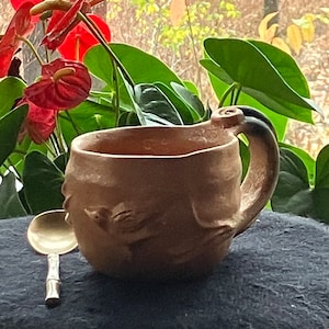 以下が含まれることがあります： ユニークな取っ手と彫刻されたデザインが施された茶色のセラミックマグカップ。マグカップは、暗い表面に銀のスプーンの隣に置かれています。