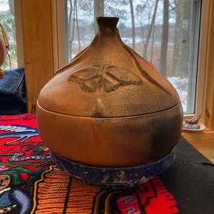 May include: A handcrafted, brown ceramic lidded jar with a bulbous body and a textured, stylized butterfly design. The jar sits on a blue and white ceramic base. The lid has a small, dark brown knob.