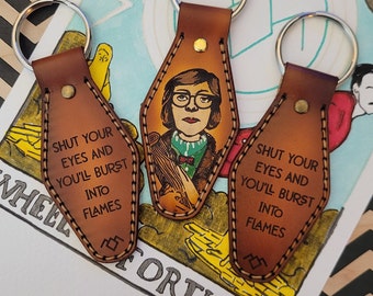 May include: Three leather keychains shaped like motel keys. Each keychain features a different image and text. The first keychain has a woman with glasses and the text "SHUT YOUR EYES AND YOU'LL BURST INTO FLAMES". The second keychain has the same text and image. The third keychain has the same text and image. The keychains are on a white background with a black and white pattern.