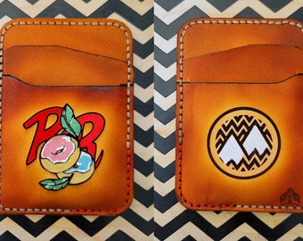 May include: Two brown leather wallets with different designs. The first wallet has a red and blue logo with the letters "PB" and a donut. The second wallet has a white mountain logo inside a circle with a black and white chevron pattern.