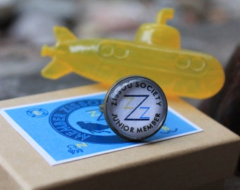 May include: A small, round, blue and white pin with the text "Zippy Society Junior Member" and a double "Z" in the center. The pin is sitting on a brown box with a blue and white label that says "Zippy Society Member". A yellow toy submarine is in the background.