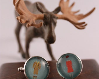 May include: A pair of cufflinks with a blue background. One cufflink features a cartoon image of a person wearing a brown and yellow scout uniform. The other cufflink features a cartoon image of a person wearing a red dress and a red hat.