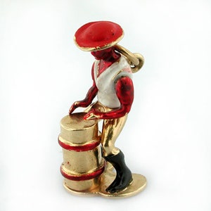 May include: A gold charm of a person wearing a red hat playing a drum. The drum is gold with red accents. The person is wearing a white shirt and red pants.