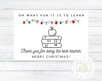 Peut inclure: Une carte blanche avec du texte noir qui dit "OH WHAT FUN IT IS TO LEARN" et "Thank you for being the best teacher MERRY CHRISTMAS!" Une illustration en noir et blanc d'une pile de livres avec une pomme sur le dessus est au centre de la carte. Une guirlande de lumières colorées est au-dessus du texte.