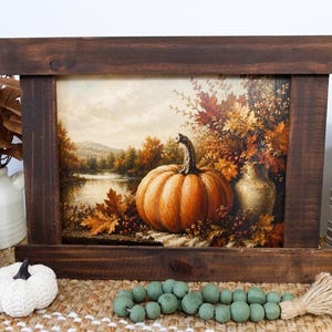 May include: A rectangular wooden frame containing a print of an autumn landscape featuring a large orange pumpkin, a ceramic vase, and colorful fall foliage by a lake. The frame is dark brown with a horizontal plank design at the top and bottom.