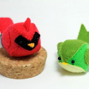 Può includere: Due ornamenti di uccelli in feltro, un cardinale rosso e un uccello verde con un becco giallo. Il cardinale è seduto su una base di sughero.