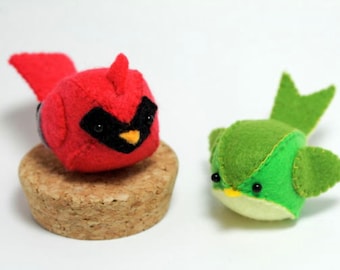 Puede incluir: Dos adornos de pájaros de fieltro, un cardenal rojo y un pájaro verde con un pico amarillo. El cardenal está sentado sobre una base de corcho.