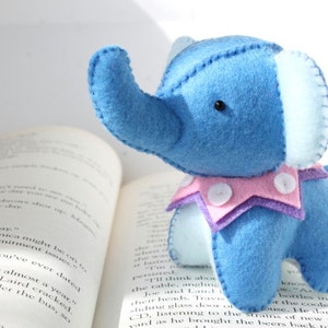 May include: A blue felt elephant toy with a pink and purple collar and white buttons. The elephant is sitting on a book.
