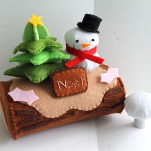 Op de afbeelding: Een kerstboomstam van vilt met een sneeuwpop, een kerstboom en een roze hulstbes. De stam is versierd met het woord "Noël" in bruin vilt.