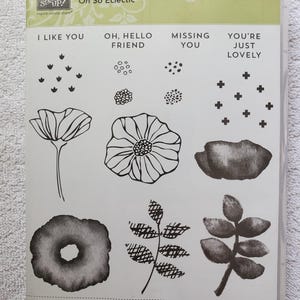 May include: A clear plastic case containing a set of 16 rubber stamps labeled Oh So Eclectic. The set features black line art designs of flowers, leaves, and abstract shapes, along with the phrases I LIKE YOU, OH, HELLO FRIEND, MISSING YOU, and YOU'RE JUST LOVELY.