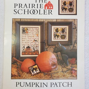 May include: A craft pattern book titled The Prairie Schooler Book No. 140 Pumpkin Patch. The cover features two framed cross-stitch designs with Halloween motifs, two orange pumpkins, a black cat figurine, and straw on a wooden surface.