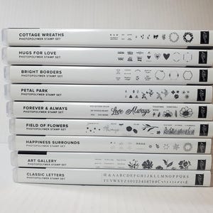 May include: A stack of clear plastic stamp sets with white labels. The labels feature titles like "Cottage Wreaths," "Hugs for Love," and "Classic Letters." Each set includes various floral and decorative designs, and letters.