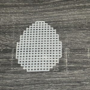 May include: A white plastic canvas sheet with a grid pattern, cut into an oval shape. The canvas is designed for cross-stitch or needlepoint projects. The background is a dark gray wood grain.