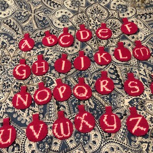 May include: A collection of red, handmade keychains, each featuring a different letter of the alphabet in white. The keychains are arranged on a patterned blue and white fabric, showcasing a personalized gift idea.