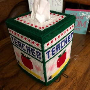 Op de afbeelding: Een groene en witte tissueboxhoes met het woord "Teacher" op de voorkant. De hoes is versierd met rode appels, een geel potlood en roze harten.
