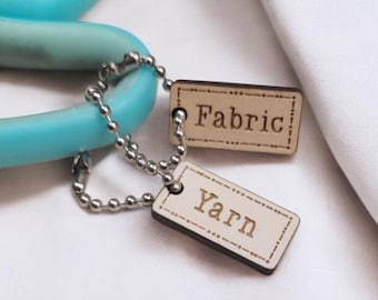 May include: Two wooden tags with string attached to a pair of blue scissors. The tags are labeled "Fabric" and "Yarn".