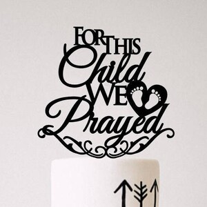 Op de afbeelding: Zwarte taarttopper met de tekst "For this child we prayed" en een hart met babyvoetjes. De taarttopper is op een witte taart met zwarte geometrische ontwerpen.