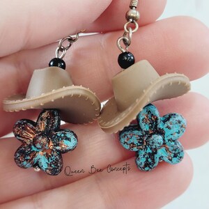 May include: A pair of dangle earrings featuring a small tan cowboy hat above a turquoise and bronze flower. The earrings have a black bead and a silver-tone hook. The text "Queen Bee Concepts" is visible.