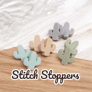 May include: A set of eight silicone cactus stitch stoppers in various shades of green, blue, and beige. The stoppers are shaped like cacti with a textured surface. The text "Stitch Stoppers" is printed on a wooden surface below the stoppers.
