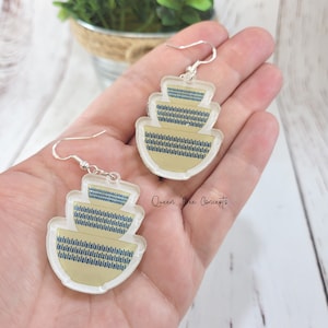 May include: A pair of clear acrylic earrings featuring a stack of three yellow and blue patterned bowls. The earrings are silver and have a hook closure. The text "Queen Bee Concepts" is visible in the image.