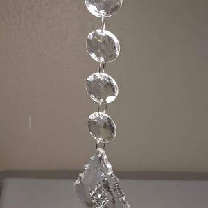 May include: A clear crystal chandelier pendant with a series of round, faceted beads connected by silver-colored metal links. The bottom features a larger, irregularly shaped crystal. The crystals reflect light.