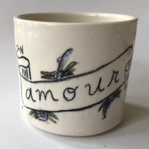May include: White ceramic mug with a black and white illustration of a banner with the word "amour" written on it. The banner is decorated with small floral designs in shades of purple, blue, and green.