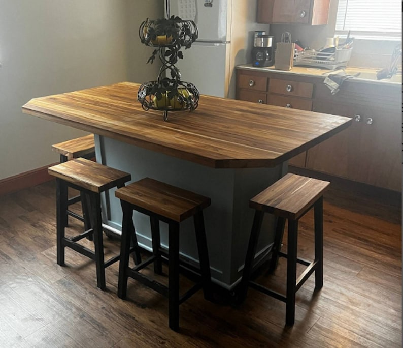 Peut inclure: Un &icirc;lot de cuisine en bois clair avec une base grise et quatre tabourets de bar en m&eacute;tal noir. L'&icirc;lot a un comptoir en bois et un bol &agrave; fruits en m&eacute;tal d&eacute;coratif.
