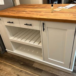 May include: White kitchen island with a wood countertop, two drawers, a cabinet door, and two open shelves. The island has black hardware.