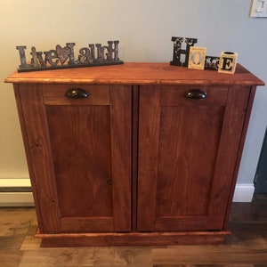 K&ouml;nnte beinhalten: Ein Holzschr&auml;nkchen mit zwei T&uuml;ren und zwei Schubladen. Der Schrank ist in einem dunklen Braunton gebeizt und hat schwarze Metallgriffe. Der Schrank ist mit den Worten "Live Laugh" und "Home" verziert.